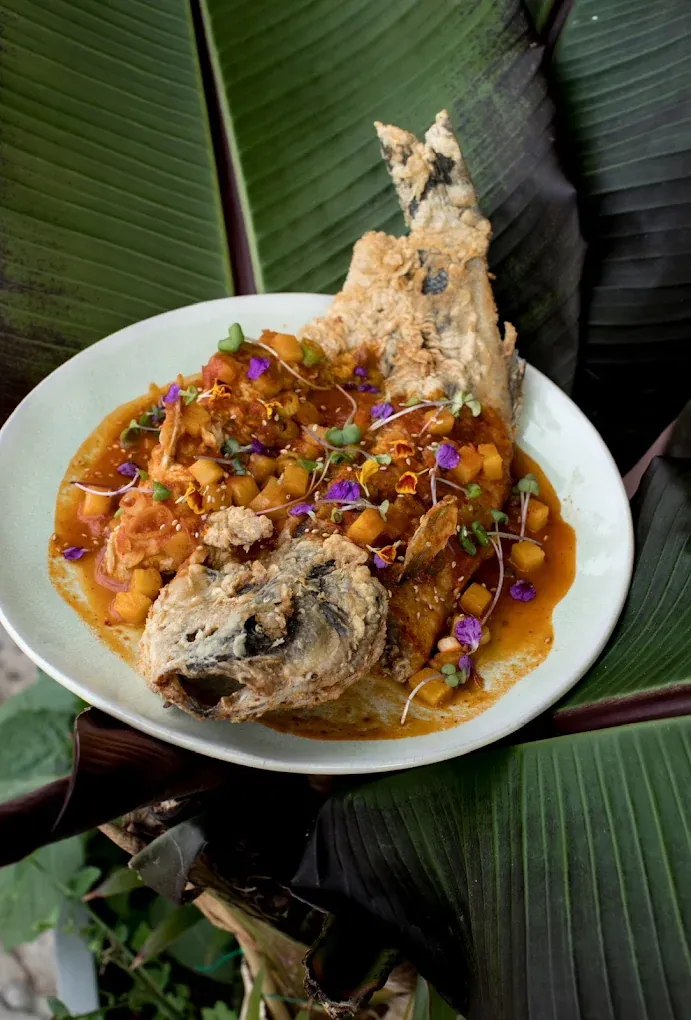 Pescado entero crujiente con salsa, dados de mango y flores comestibles, sobre hojas de platanera